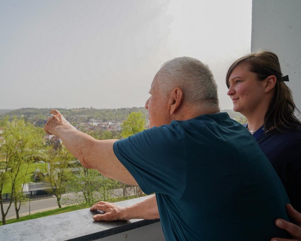 Älterer Herr zeigt vom Balkon über die Stadt, eine Pflegerin steht neben ihm