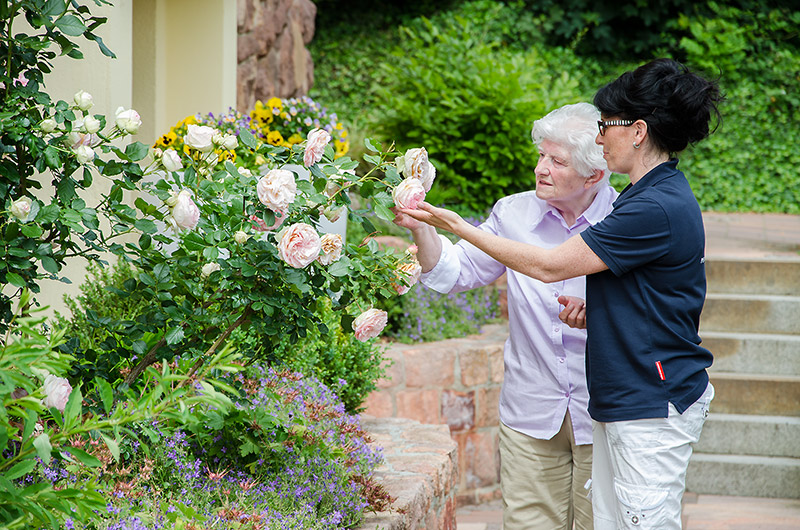Eine Pflegerin und eine ältere Dame betrachten gemeinsam Rosen im Garten