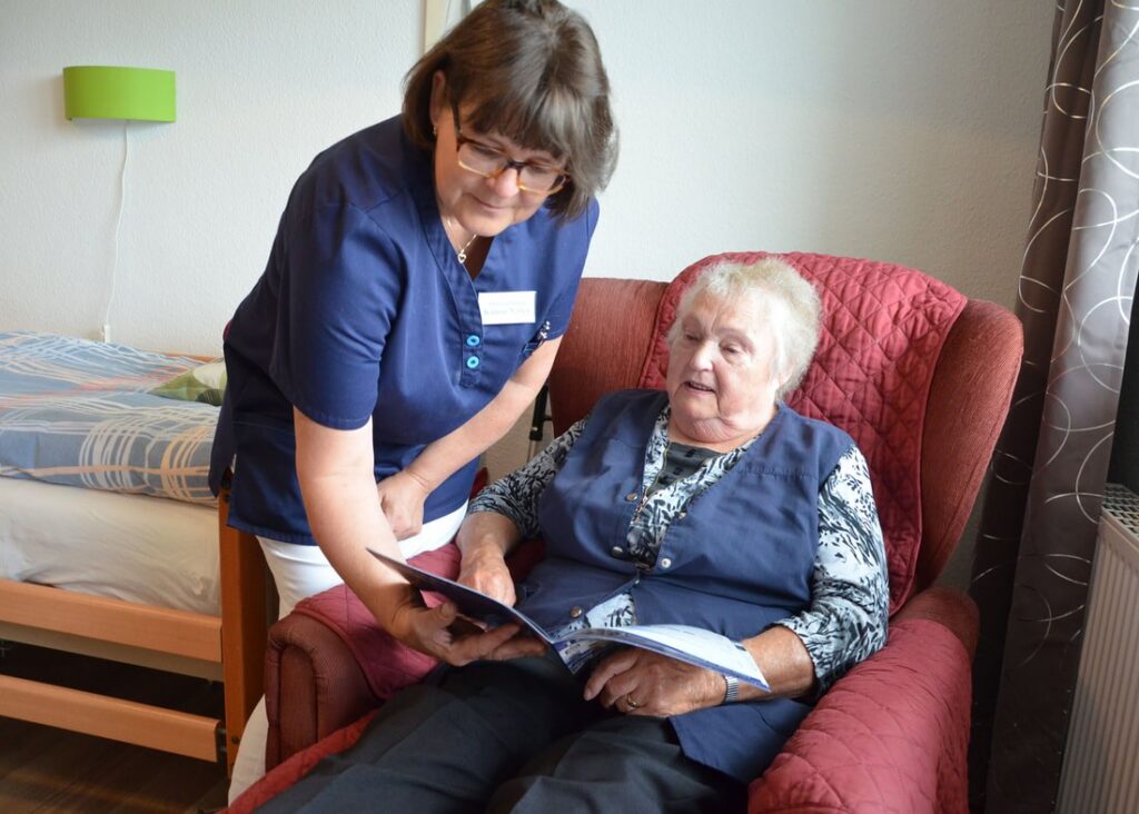 Pflegerin zeigt einer Seniorin im Sessel ihrer Wohnung eine Broschüre