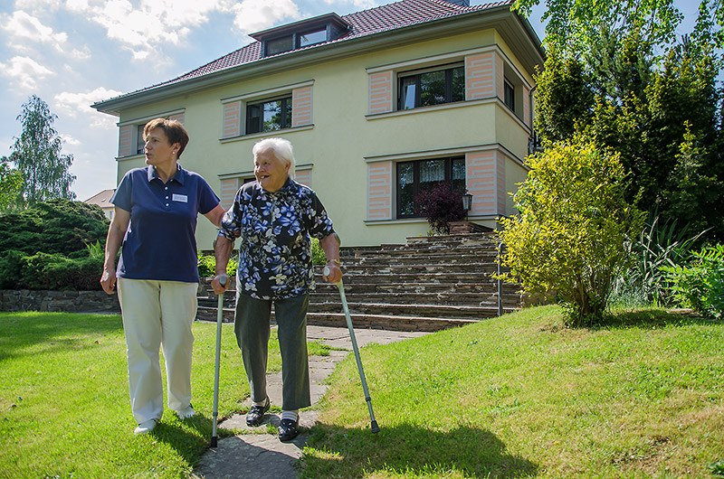 Pflegerin begleitet eine Seniorin mit Gehstützen über den Rasen vor der Demenz-Wohngemeinschaft in Roßwein
