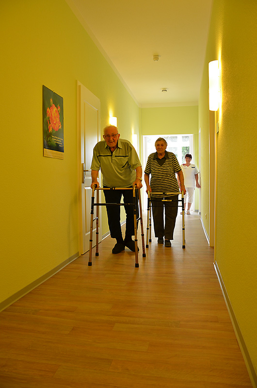 Zwei ältere Menschen gehen mit Rollatoren durch einen gelb gestrichenen Flur