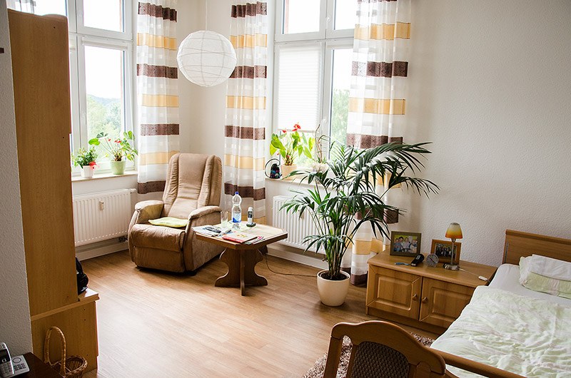 Helles Zimmer im Betreuten Wohnen mit eigenen Möbeln, Sessel, Pflanzen und Bett