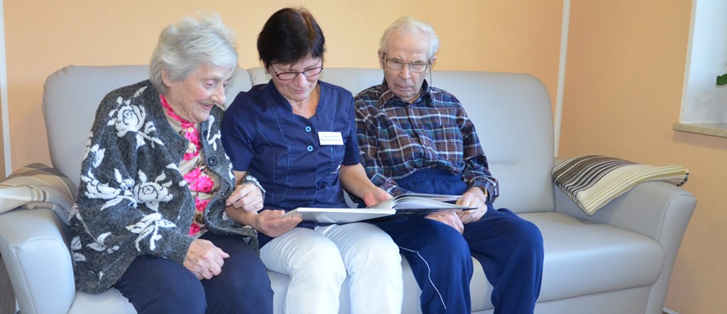 Pflegerin sitzt zwischen einer Bewohnerin und einem Bewohner auf dem Sofa und blättert mit ihnen in einem Buch