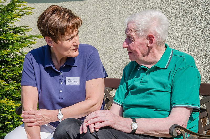 Pflegerin und älterer Herr sitzen in der Sonne auf einer Bank und unterhalten sich vertraut