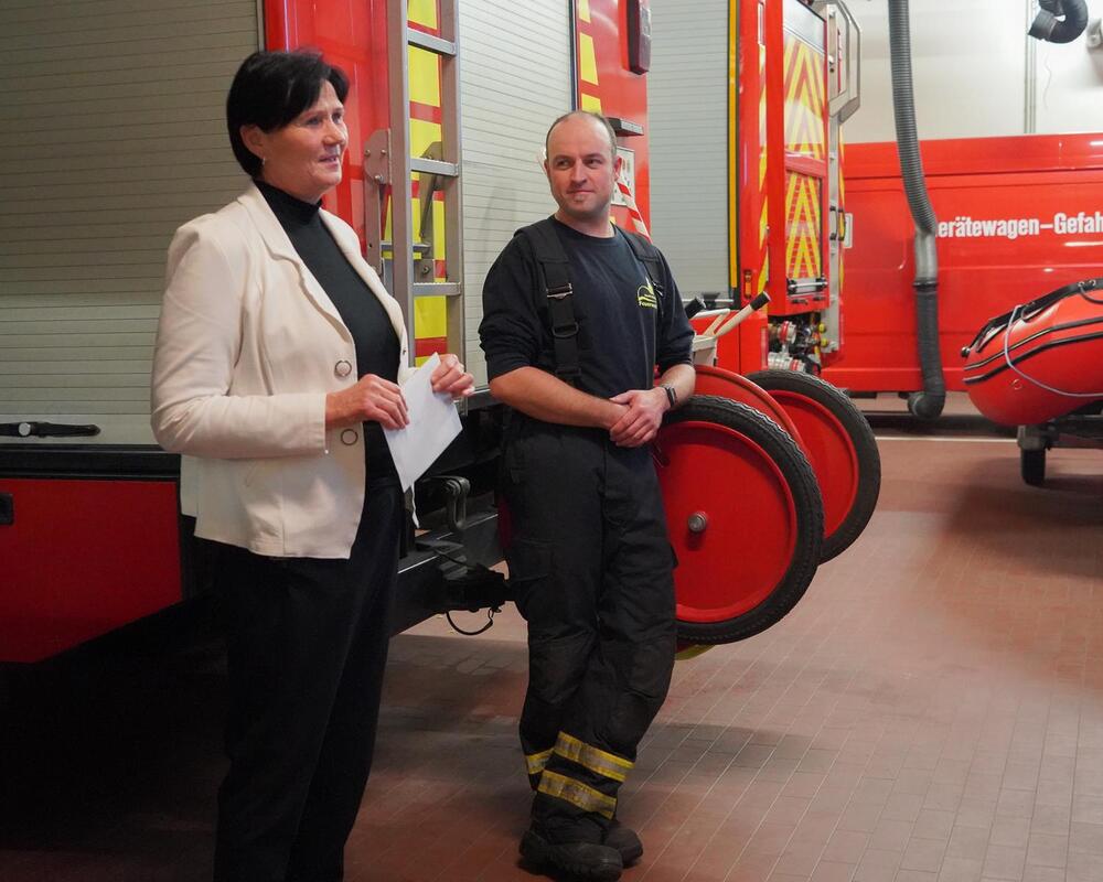 Spendenübergabe in der Fahrzeughalle der Döbelner Feuerwehr: eine Frau im hellen Blazer neben einem Feuerwehrmann, dahinter ein Löschfahrzeug