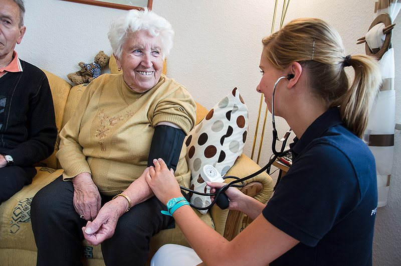 Pflegerin misst einer Seniorin im Sessel den Blutdruck
