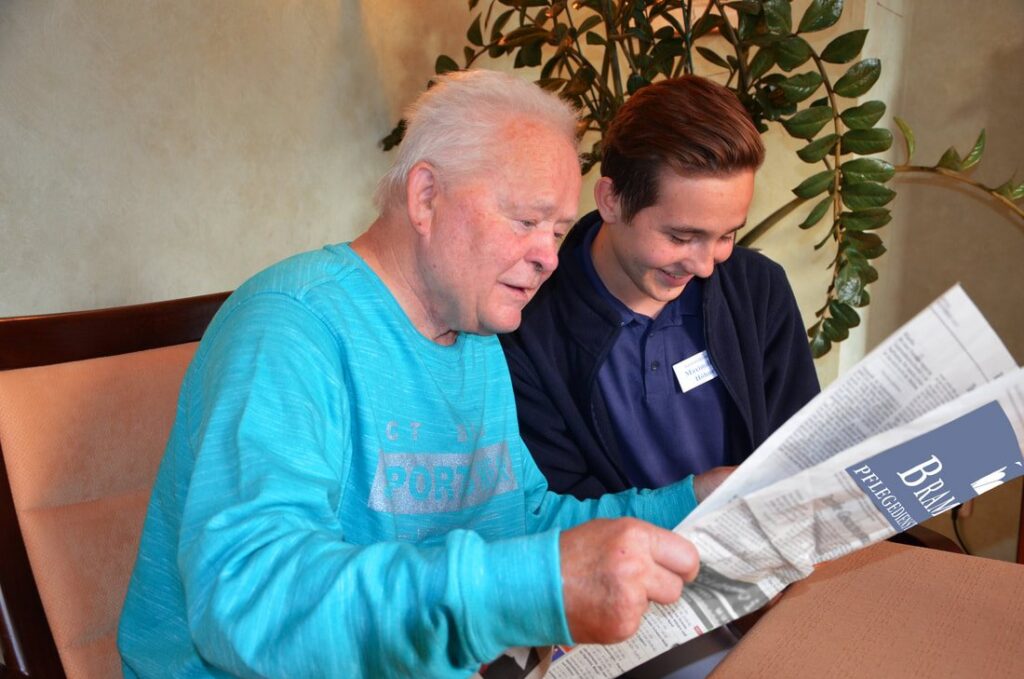 Junger Pflegeschüler liest mit einem älteren Herrn gemeinsam Zeitung
