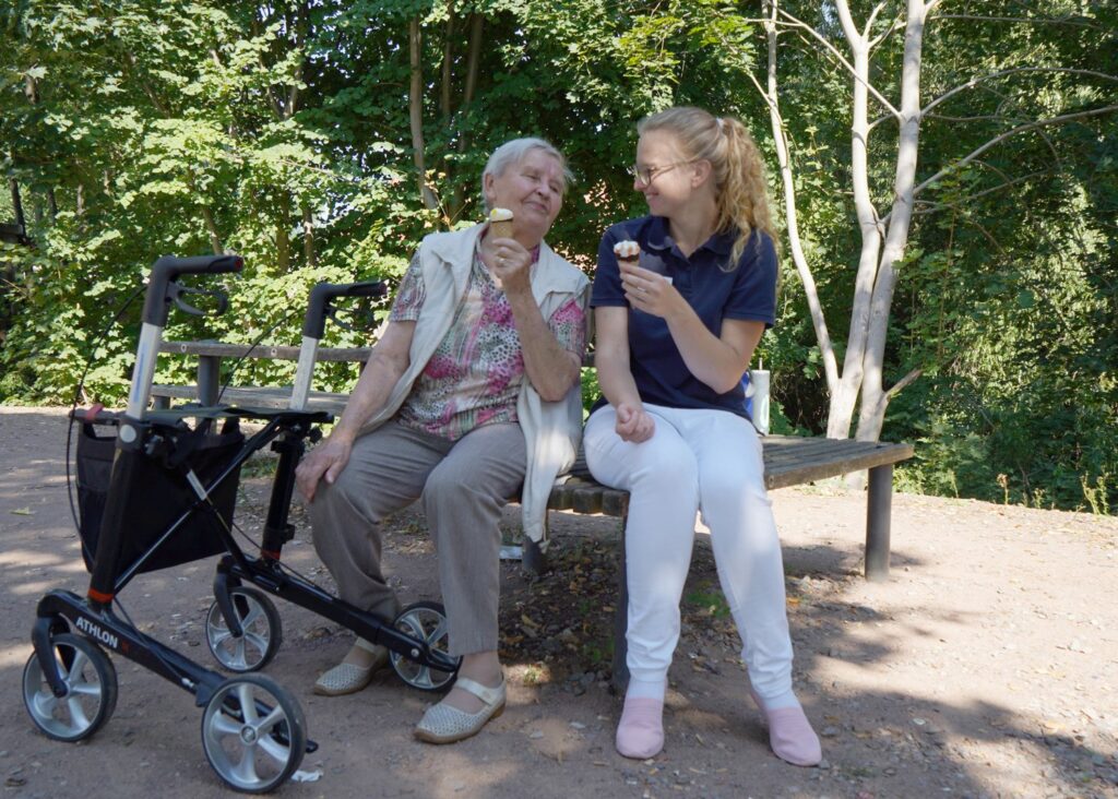 Junge Pflegerin und Seniorin essen gemeinsam ein Eis auf einer Parkbank