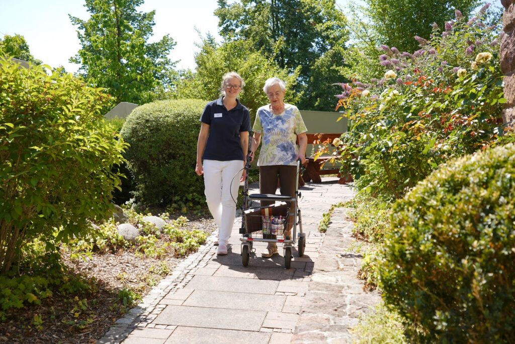 Pflegerin begleitet eine Seniorin mit Rollator auf einem Gartenweg