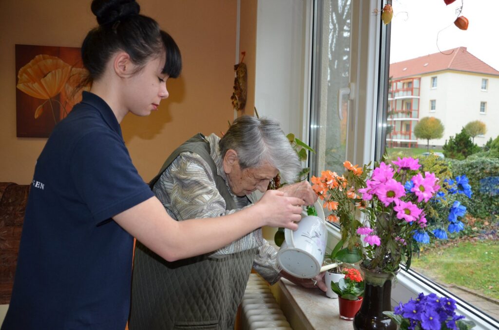 Junge Pflegekraft gießt mit einer Seniorin Blumen am Fenster