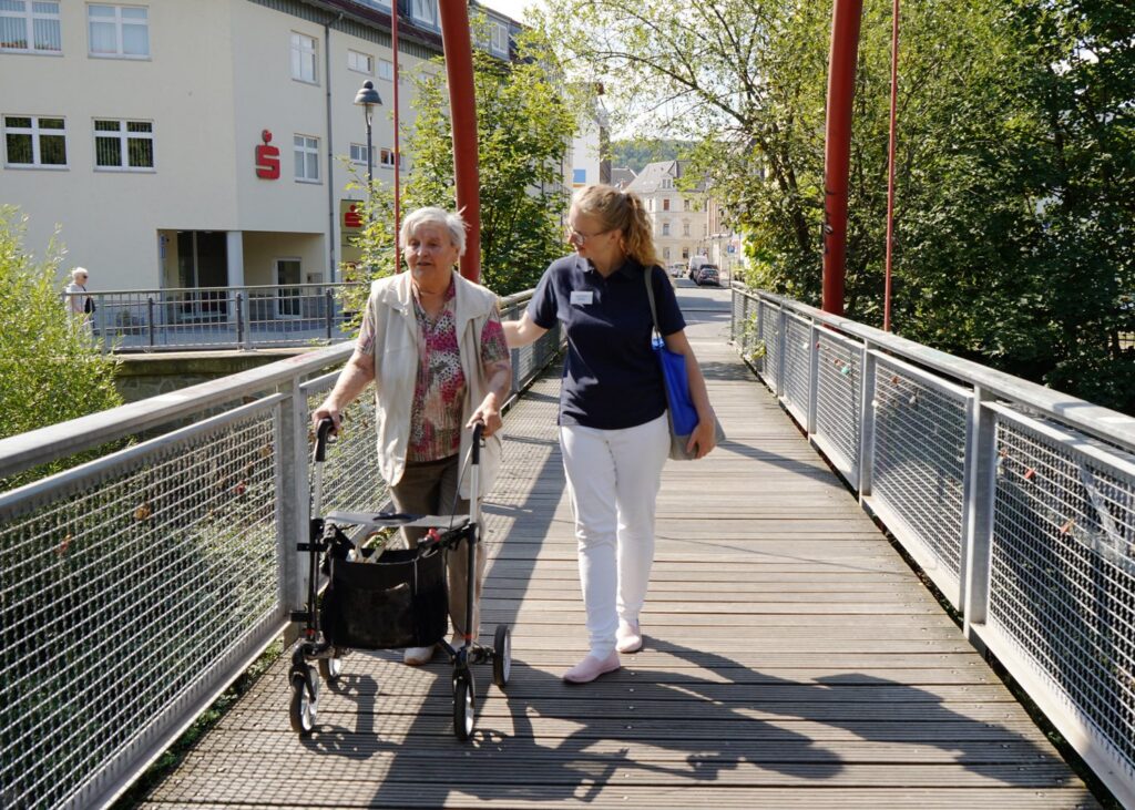 Junge Pflegekraft begleitet eine Seniorin mit Rollator über eine Brücke