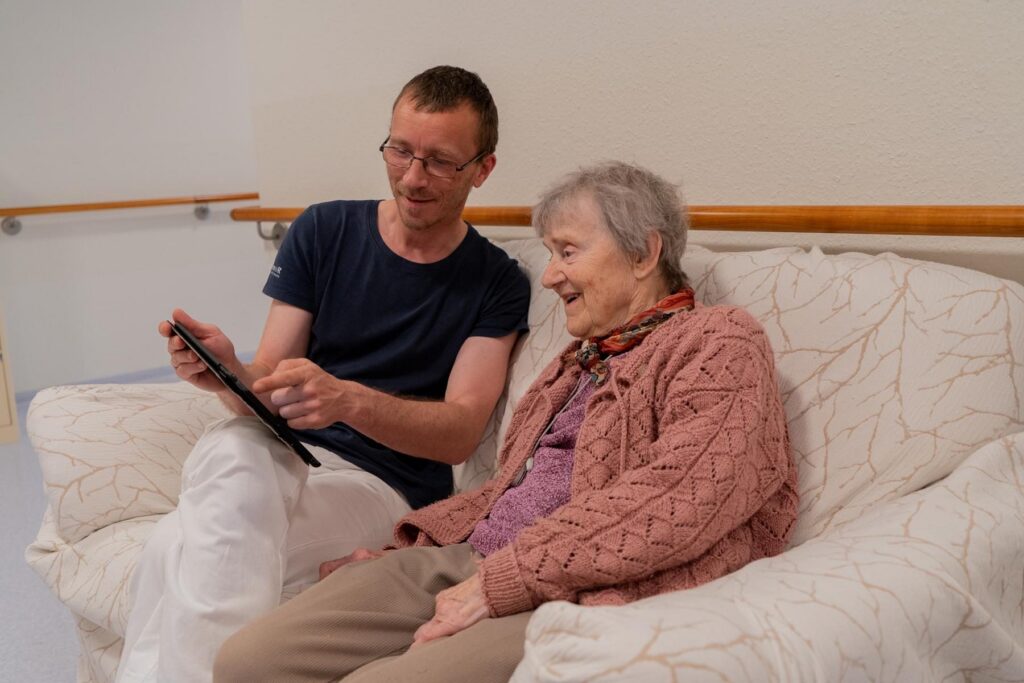 Pfleger und Seniorin sitzen auf einem Sofa und schauen gemeinsam auf ein Tablet