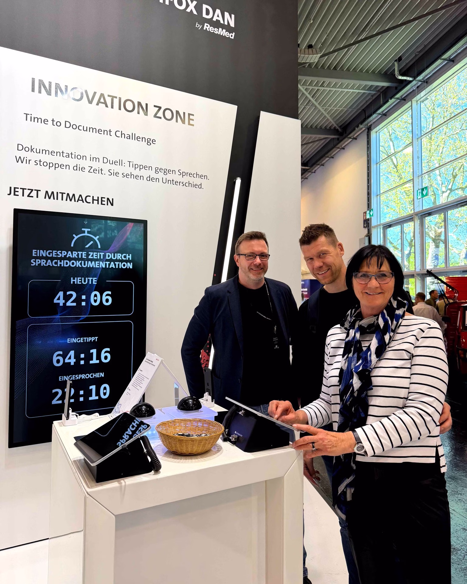 Drei Mitarbeitende am Messestand von MEDIFOX DAN vor einer Anzeige zur eingesparten Dokumentationszeit