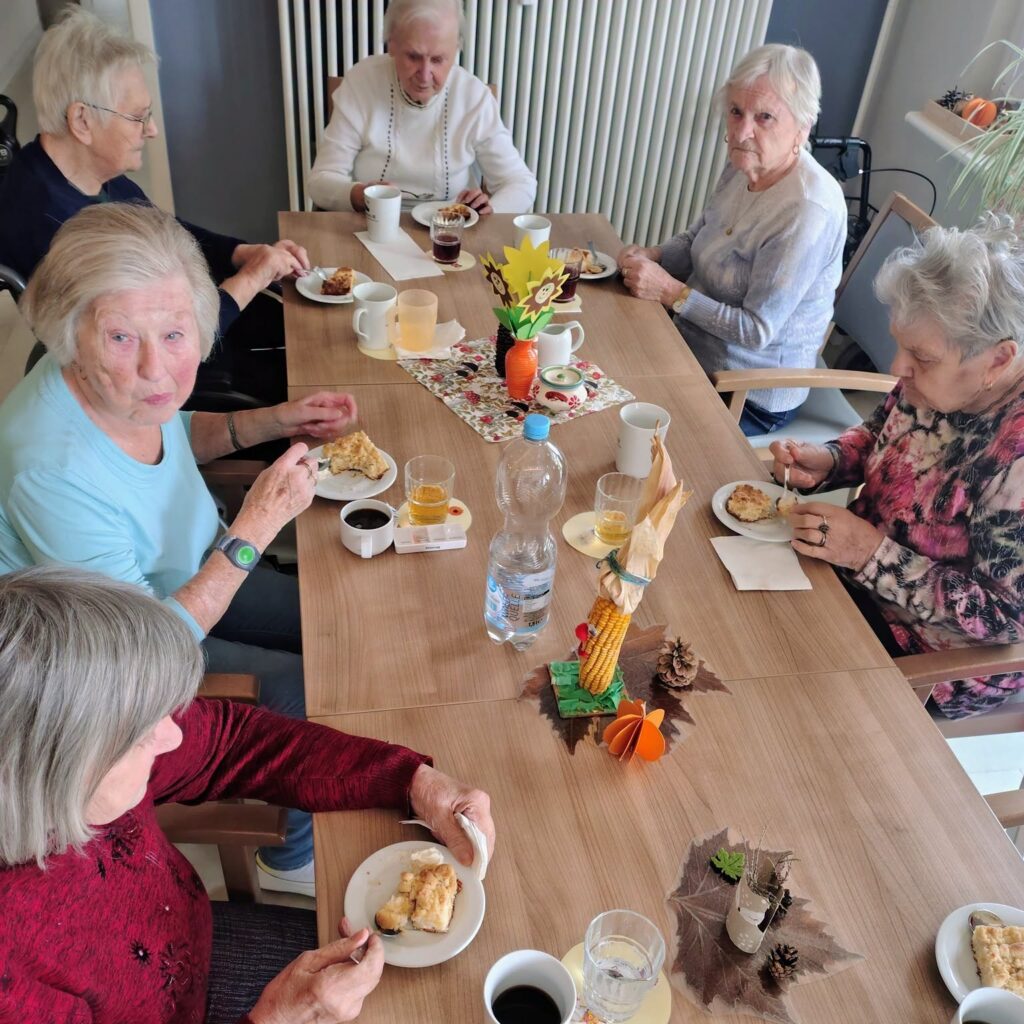 Seniorinnen sitzen an einem langen Tisch mit Herbstdekoration und essen Apfelkuchen mit Streuseln zum Kaffee