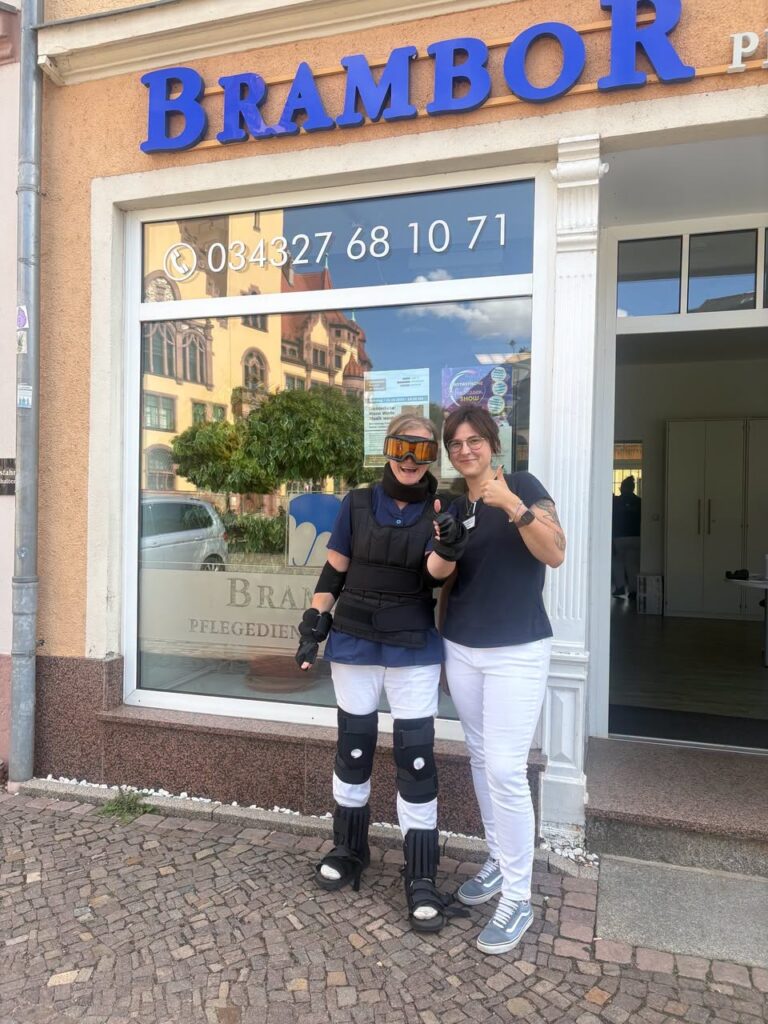 Eine Mitarbeiterin im Altersimulationsanzug mit Schutzbrille, Weste und Gelenkstützen steht neben einer Kollegin vor dem Brambor-Büro in Waldheim