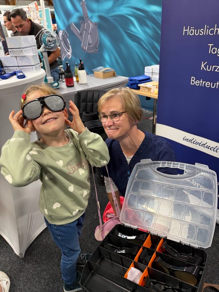 Ein Mädchen probiert am Brambor-Stand eine Simulationsbrille aus, neben ihr lächelt eine Mitarbeiterin, vor ihnen steht ein Koffer mit weiteren Brillen