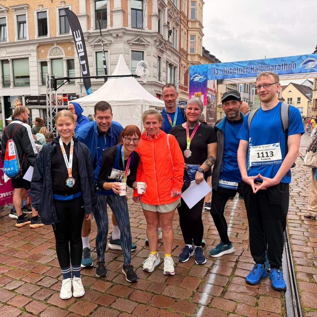 Eine Gruppe Läuferinnen und Läufer mit Medaillen steht bei Regen auf dem Pflaster in der Döbelner Innenstadt vor dem Zielbogen