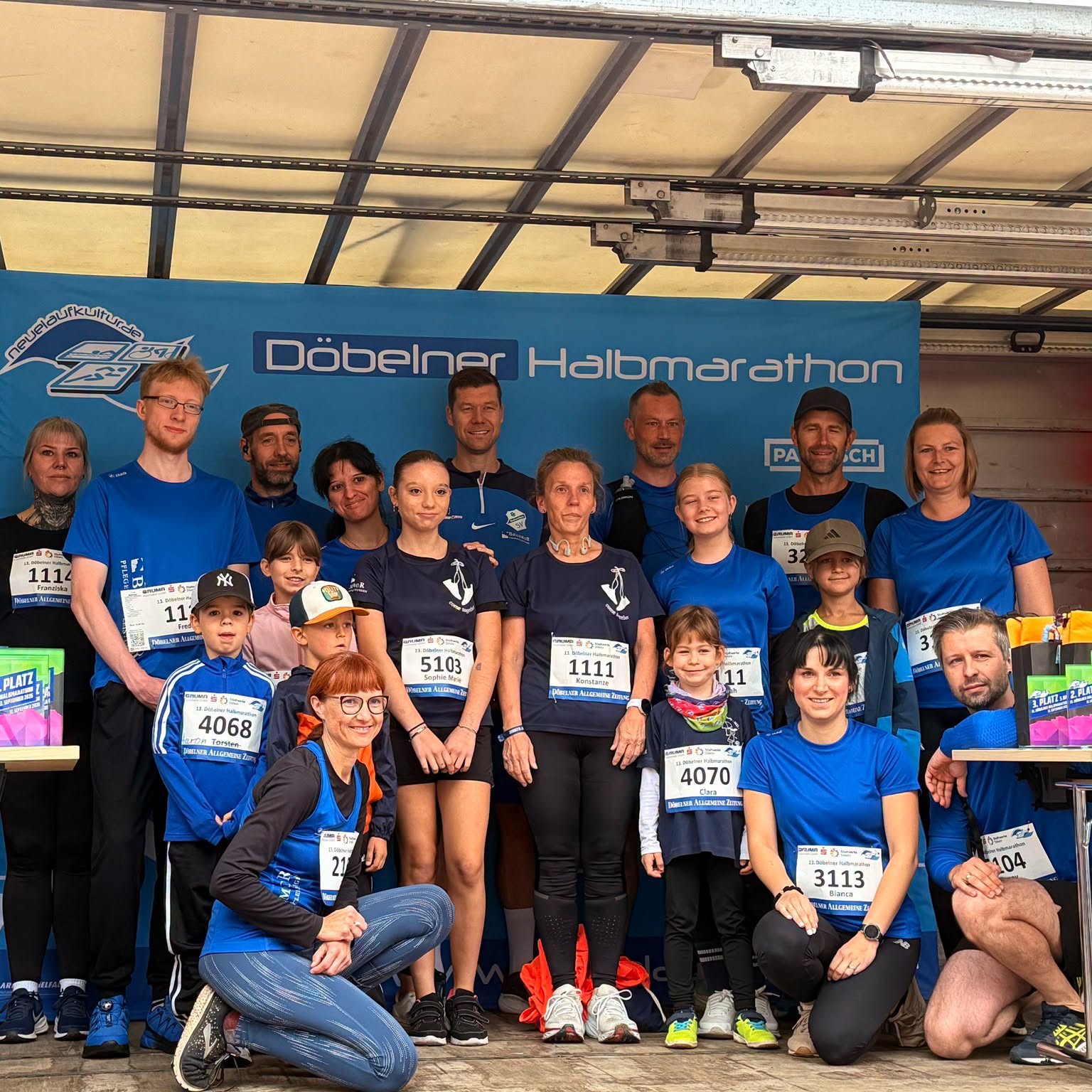 Das Brambor-Läuferteam mit Erwachsenen und Kindern steht in blauen Shirts mit Startnummern vor dem Banner des Döbelner Halbmarathons