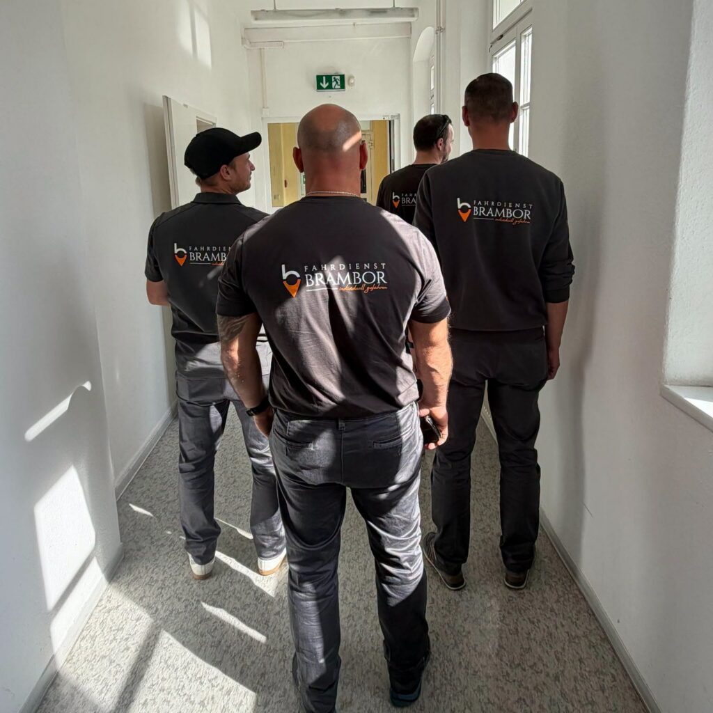 Vier Mitarbeiter des Fahrdienstes in dunklen Shirts mit dem Logo „Fahrdienst Brambor“ gehen von hinten gesehen durch den Flur