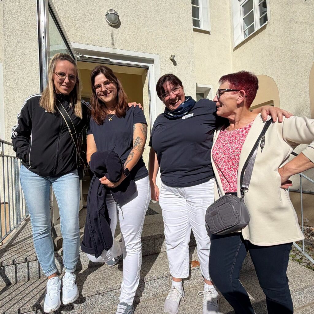 Vier lachende Kolleginnen stehen untergehakt vor dem Eingang des Gebäudes