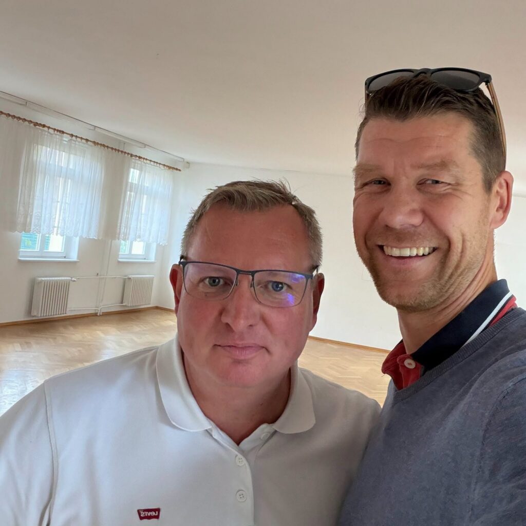 Zwei Männer machen ein Selfie in einem leeren, hellen Saal mit Parkettboden