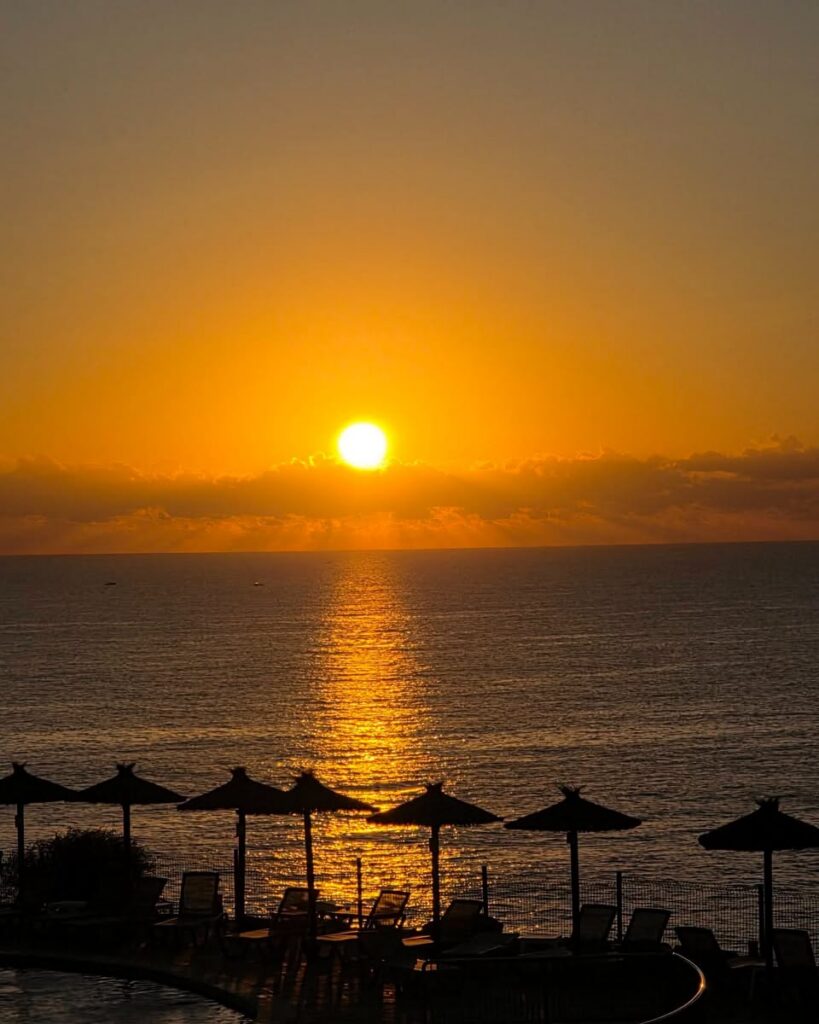 Sonnenuntergang über dem Meer, im Vordergrund die Strohschirme einer Hotelanlage