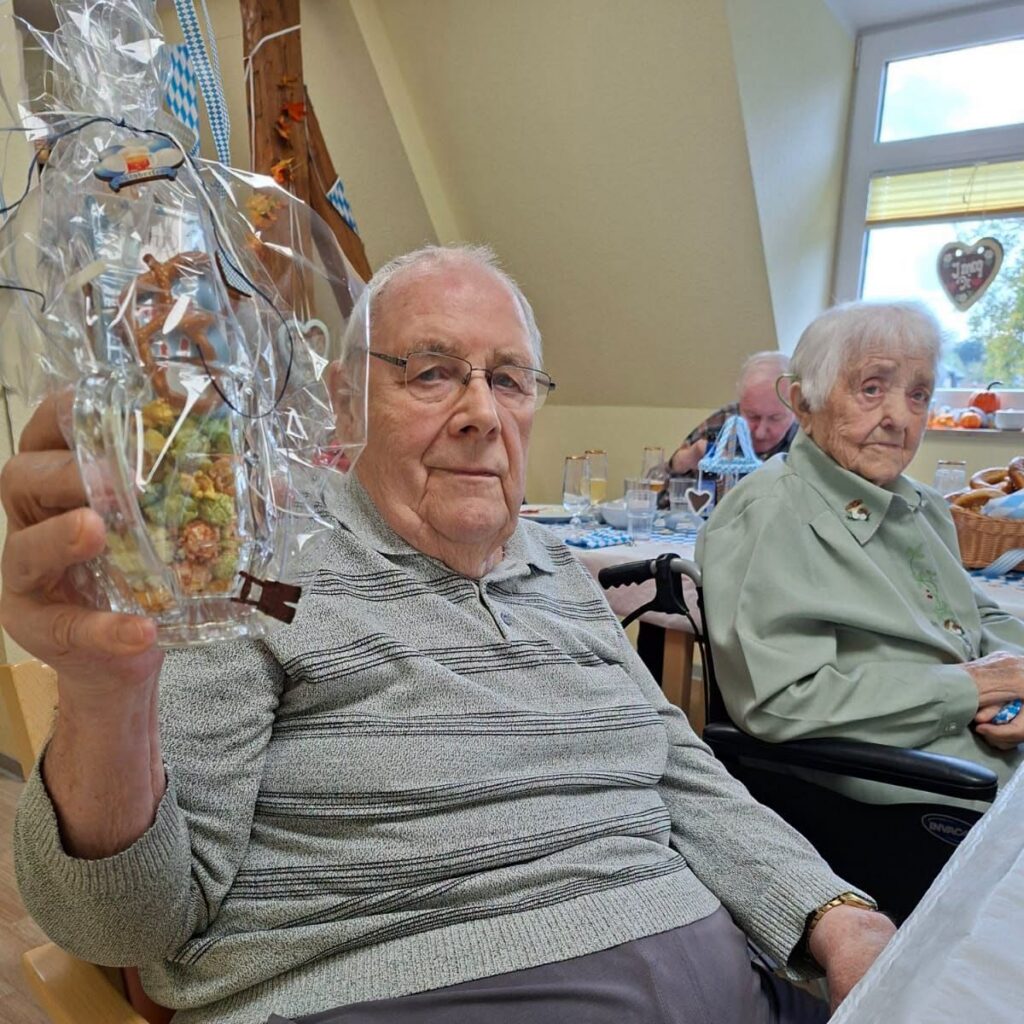 Ein Bewohner mit Brille hält ein Glas mit Knabbereien in Folie hoch, daneben sitzt eine Bewohnerin im Rollstuhl
