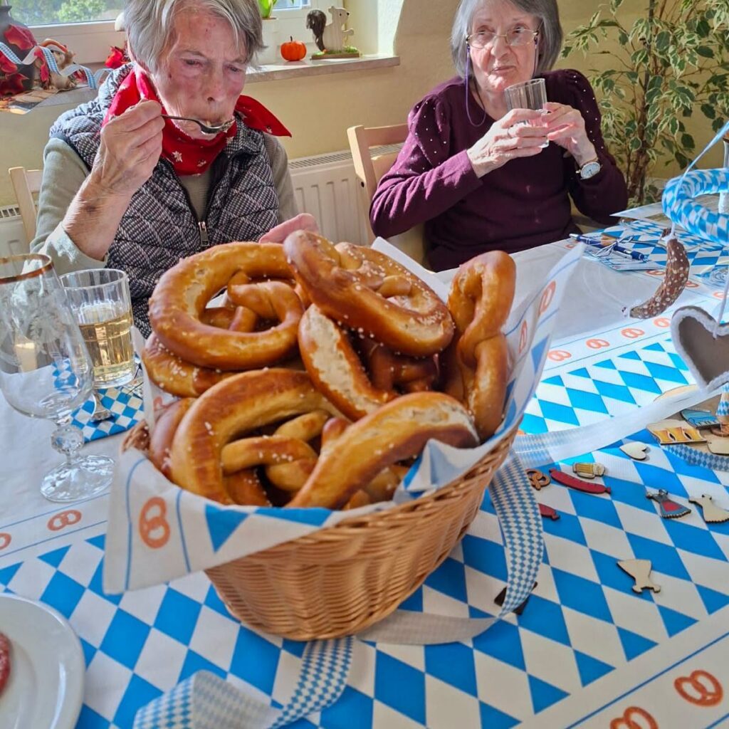 Korb mit frischen Brezeln auf einer blau-weißen Tischdecke, dahinter zwei Bewohnerinnen beim Essen und Trinken