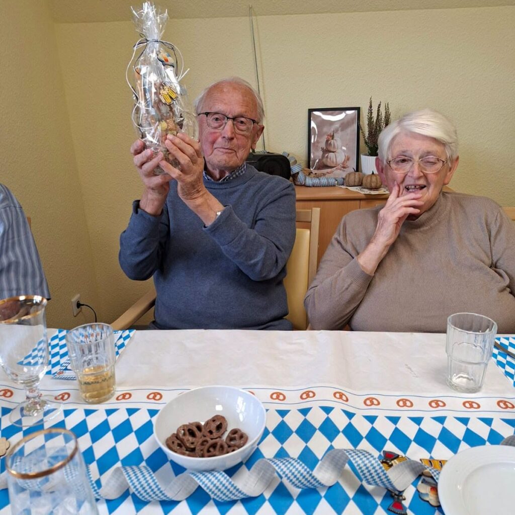 Ein Bewohner hält ein Geschenkglas hoch, neben ihm lacht eine Bewohnerin; auf dem Tisch stehen Brezeln und blau-weiße Dekoration