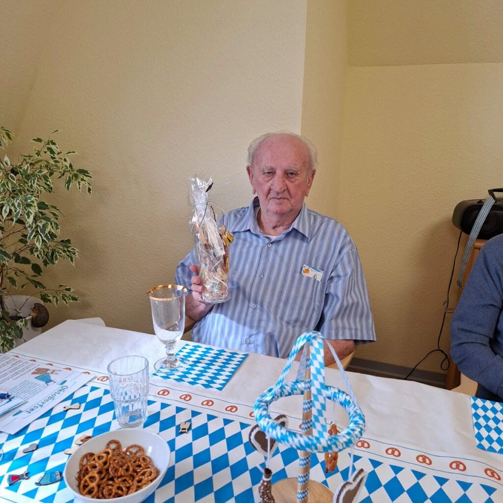 Ein Bewohner hält am festlich gedeckten Tisch ein Geschenkglas, vorn stehen ein Maibaum aus blau-weißen Bändern und eine Schale mit Salzbrezeln