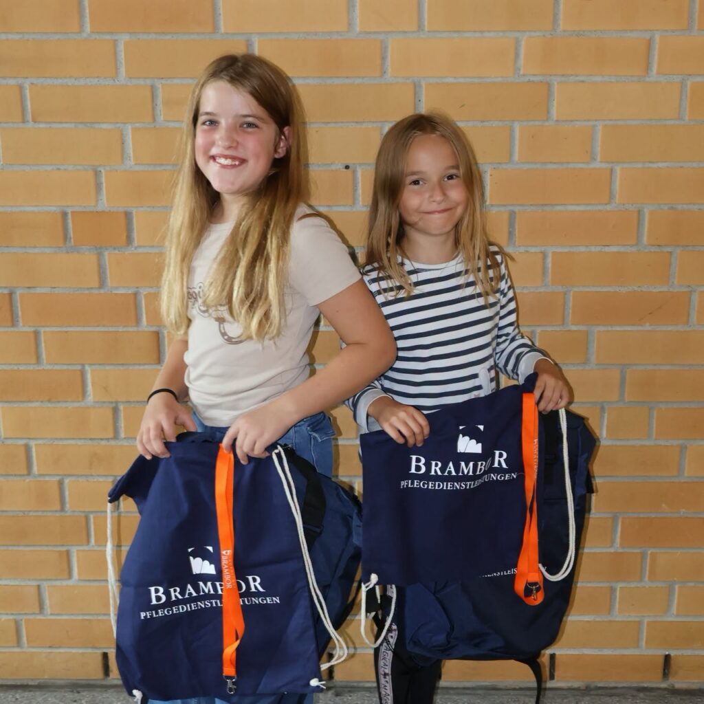 Zwei Mädchen halten vor einer Backsteinwand dunkelblaue Brambor-Turnbeutel mit orangefarbenen Bändern