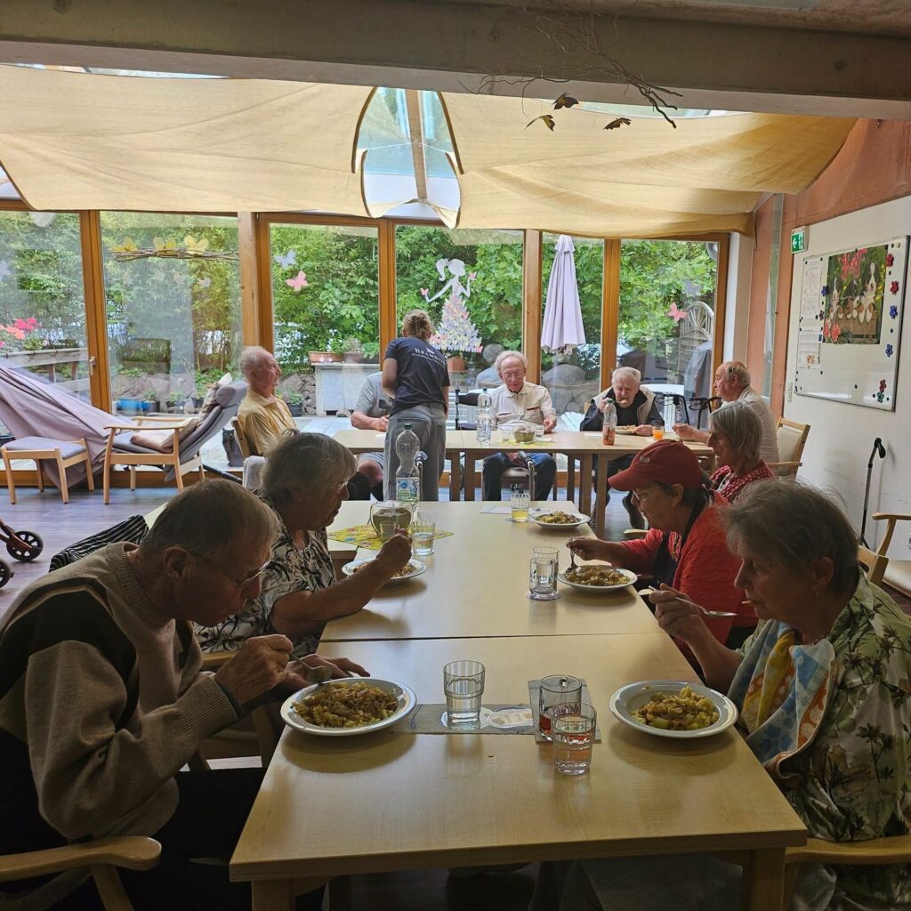 Gäste der Tagespflege sitzen an einem langen Tisch im hellen Wintergarten beim gemeinsamen Essen