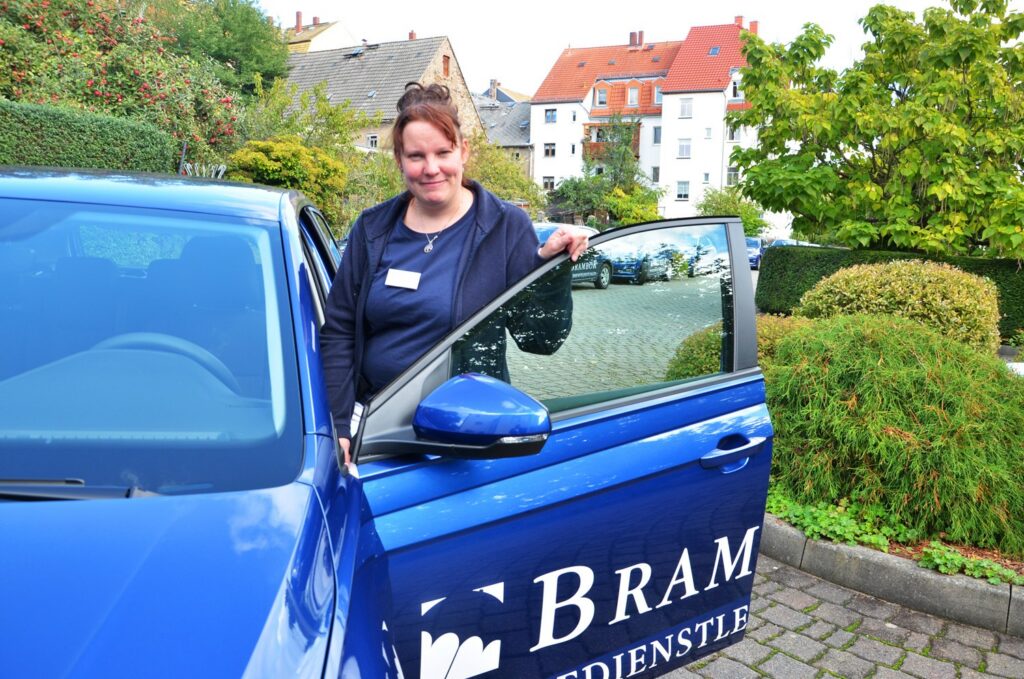 Lächelnde Pflegerin steht in Roßwein an der offenen Tür eines blauen Brambor-Dienstwagens