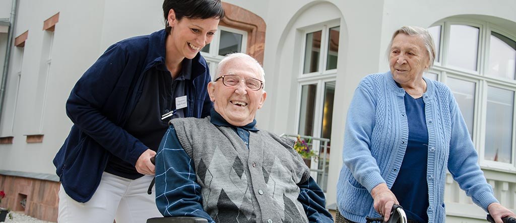 Pflegerin begrüßt einen lachenden Herrn im Rollstuhl, daneben eine Seniorin mit Rollator