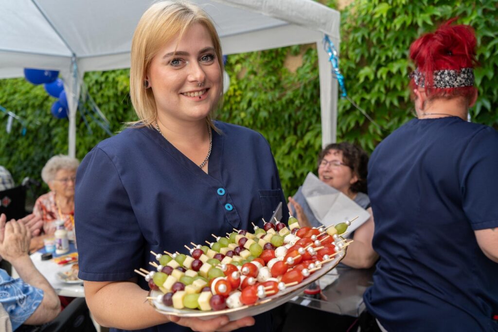Lächelnde Pflegerin serviert beim Sommerfest in Waldheim eine Platte mit Obstspießen