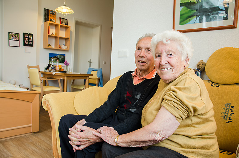 Ein älteres Paar sitzt Hand in Hand auf einem gelben Sofa und lächelt