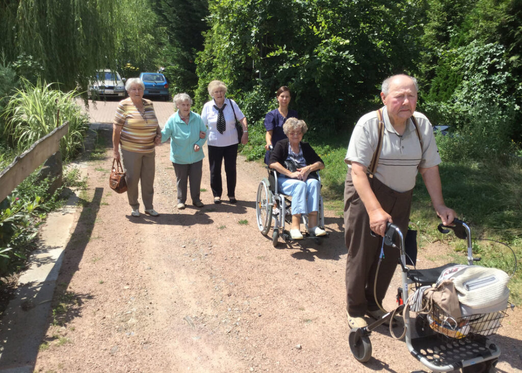 Gäste der Tagespflege mit Rollator und Rollstuhl bei einem Spaziergang auf einem Waldweg, begleitet von Mitarbeiterinnen