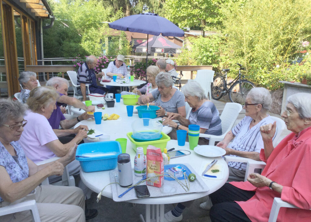 Gäste der Tagespflege „Zur Sonne“ sitzen an einem sonnigen Tag an gedeckten Tischen auf der Terrasse im Garten