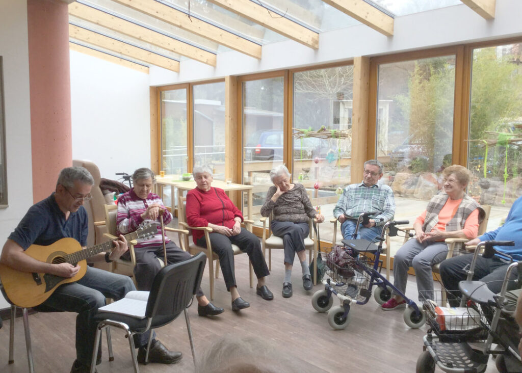 Gäste sitzen im Kreis im hellen Wintergarten der Tagespflege und hören einem jungen Mann mit Gitarre zu