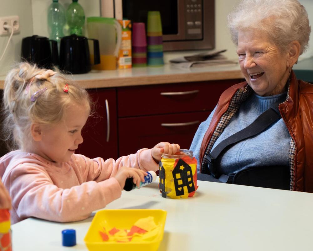 Lachende Seniorin bastelt mit einem kleinen Mädchen ein buntes Windlicht