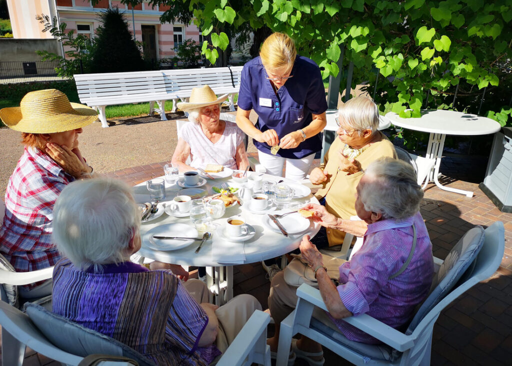Gäste sitzen mit einer Mitarbeiterin an gedeckten Tischen im Park der Villa „Zum Rüderpark“
