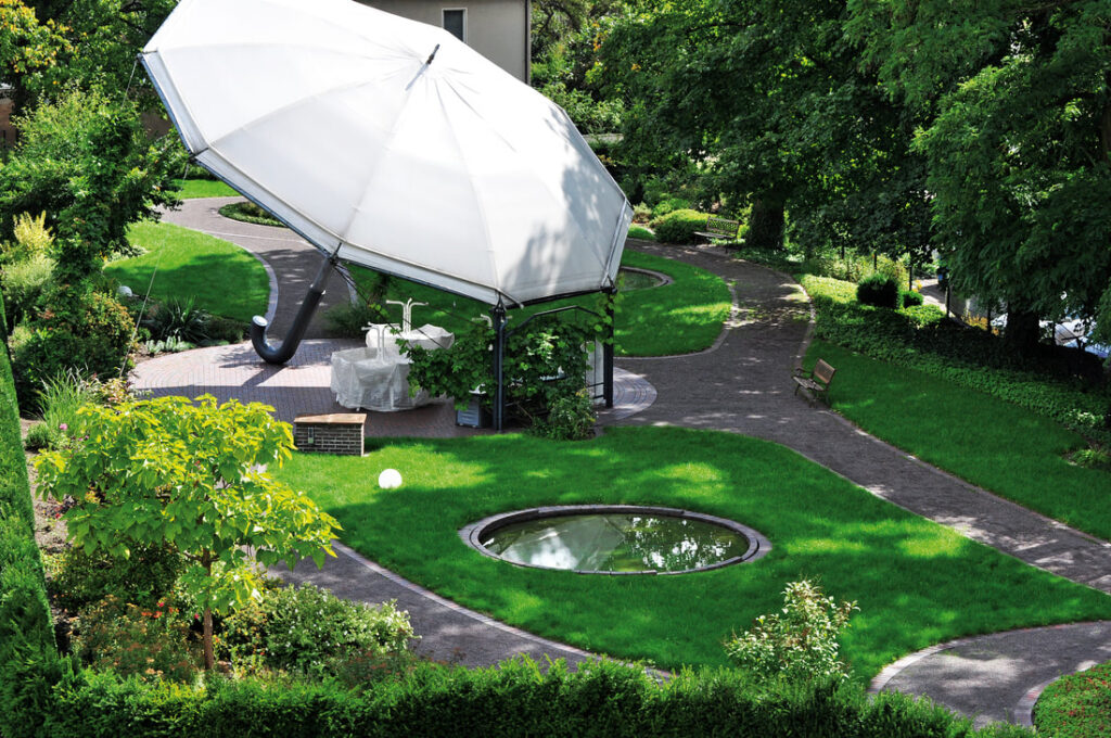 Park der Villa „Zum Rüderpark“ von oben mit großem weißem Sonnenschirm, geschwungenen Wegen und rundem Wasserbecken