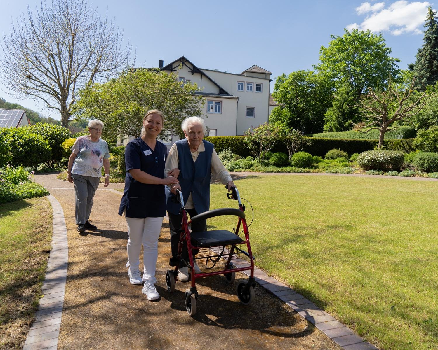Zwei Pflegerinnen begleiten zwei Seniorinnen mit Rollator bei einem Spaziergang durch den Park der Villa „Zum Rüderpark“